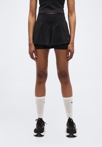 Άτομο που φοράει μαύρα στρωμένα σορτς, λευκές κάλτσες μέχρι τη μέση της γάμπας με λογότυπο και μαύρα αθλητικά παπούτσια, στέκεται μπροστά σε λευκό απλό φόντο.