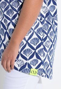 Chemise bleu et blanche à motifs de coquillage, avec une étiquette latérale en néon jaune, associée à un pantalon blanc. Texture en coton lisse.
