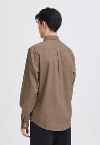 Chemise à manches longues boutonnée, de couleur marron, en tissu doux avec un ourlet arrondi, un pli au dos et des poignets ajustables. Texture lisse.