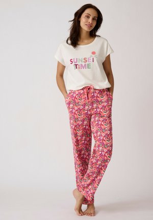 Vrouw draagt een wit T-shirt met de tekst "SUNSET TIME" en kleurrijke roze broek met bloemenpatroon, staat blootsvoets op een effen achtergrond.