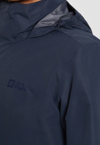 Veste d'extérieur bleu marine avec une capuche partiellement visible et un logo subtilement texturé sur le haut de la poitrine.