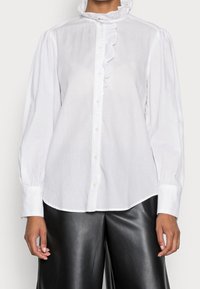 Blusa bianca a maniche lunghe con colletto arricciato e placca frontale. Realizzata in tessuto leggero; presenta maniche voluminose e dettagli con bottoni.