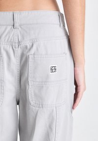Los pantalones cargo de color gris claro presentan una cintura alta, dos bolsillos traseros y un pequeño parche con logo en el bolsillo derecho. Textura de tela suave y duradera.