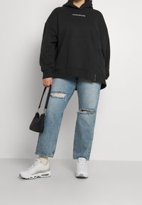 Felpa nera oversize con logo bianco, jeans strappati di un blu chiaro, sneakers bianche con dettagli grigi e una piccola borsa nera.