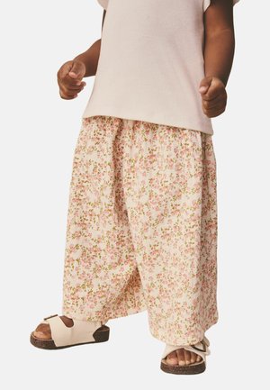 Barn iført brede bukser med blomsterprint, beige ærmeløs top og beige sandaler med korksåler, stående med knyttede næver.