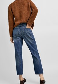 Jeans in denim blu con una forma leggermente affusolata, dotati di due tasche posteriori e un lavaggio medio. Indossati con un maglione marrone strutturato.