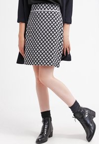 Polvella musta, joka ulottuu polveen ja jossa on valkoinen timanttikuvio, yhdistettynä mustiin nauhakengissä nilkkapohjassa ja tummiin sukkiin. Täydellisen sileä pinta ja istuva muoto.