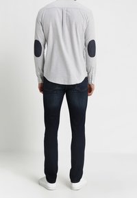 Szara koszula na guziki z ciemnymi łatami na łokciach, zestawiona z ciemnymi dżinsami i białymi trampkami. Prosty fason z detalem na tylnym karczku.