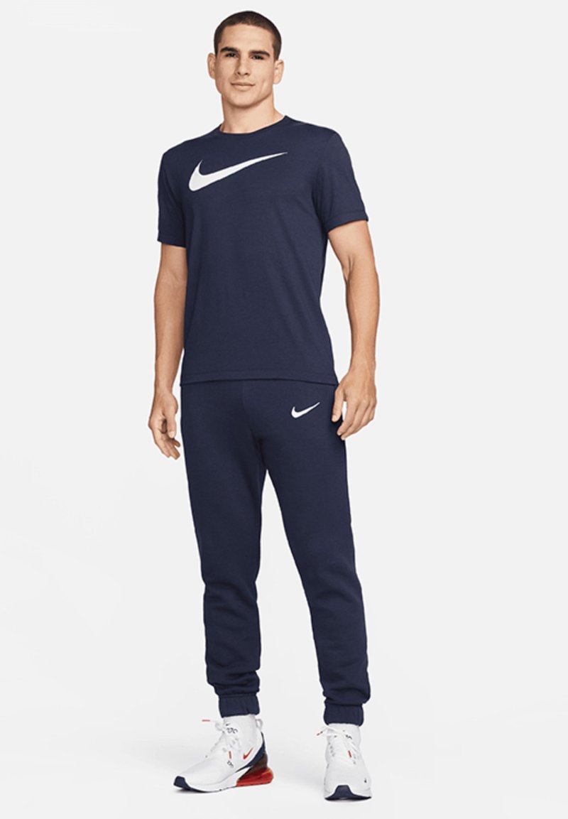 Navy Nike-T-Shirt mit weißem Logo, kombiniert mit marineblauen Jogginghosen mit weißem Logo und weißen Sportschuhen mit roten Akzenten.