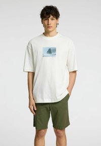 Vit bomull t-shirt med ett stort grafiskt tryck av en klippvägg över vatten. Avslappnad passform, kortärmad, ihop med olivgröna shorts.