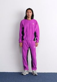 Violet sportinis megztinis su atitinkančiomis kelnių, juodais šoniniais intarpais. Ant krūtinės ir ant kojos yra mažas trikampis logotipas.