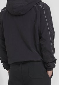 Man wearing black hooded jacket with light piping on sleeves and black pants with hand in pocket, shown from the back.