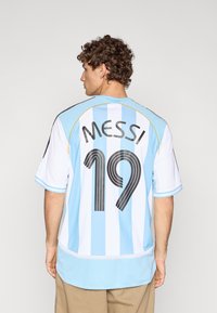 Αργεντινική φανέλα ποδοσφαίρου με κάθετες ρίγες σε ανοιχτό γαλάζιο και λευκό. Φέρει μαύρο κείμενο "MESSI" και αριθμό "19" στην πλάτη.