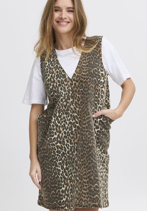 Kvinde smiler iført en ærmeløs vest med leopardprint over en hvid kortærmet skjorte, den ene hånd i vestens lomme, mod en ensfarvet baggrund.