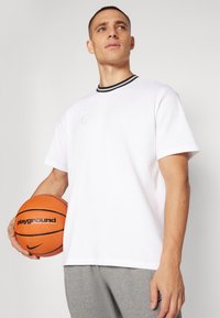 Hvit T-skjorte med kort erm og svart ribbekrage, brodert logo og brukt sammen med grå shorts, mens man holder en oransje basketball.