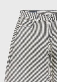 Gros plan d’un pantalon à rayures verticales noires et blanches montrant la ceinture, le bouton, la passants de ceinture et les détails de la poche avant.