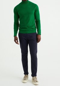Pull col roulé vert en maille légère à manches longues, associé à un pantalon bleu marine foncé et des baskets beiges.