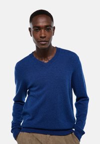 Homme portant un pull en V bleu et un pantalon marron clair, se tenant les mains dans les poches devant un fond clair uniforme.