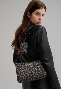 Borsa a fantasia blu navy e beige con tasca con zip, tracolla rimovibile e dettagli in argento. Texture liscia e forma arrotondata. Logo del marchio visibile.