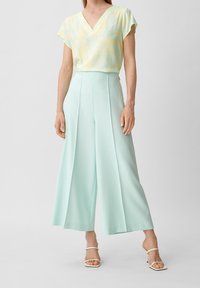 Ljusgul blommönstrad V-ringad topp i kombination med högmidjade, pastellmintfärgade vida byxor. Modellen bär vita sandaletter med remmar.