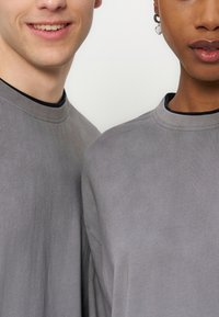 Δύο άτομα φορούν ταυτόσημες γκρι μπλούζες με στρογγυλή λαιμόκοψη, σε λευκό απλό φόντο, το ένα χαμογελάει, το άλλο φοράει σκουλαρίκι σε σχήμα καρδιάς.