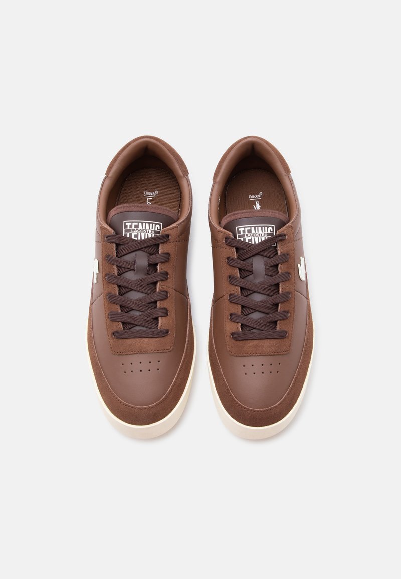 Een paar bruine leren en suède sneakers met donkerbruine veters en witte rubberen zolen op een witte achtergrond.