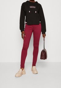 Zwarte cropped hoodie met roze logo, rode skinny jeans, beige platform schoenen, en een bordeaux handtas in de hand. Eenvoudig ontwerp, casual pasvorm.