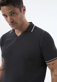 Camisa polo negra con textura y cuello en V, con franjas blancas contrastantes en el collar. Presenta mangas cortas y un diseño ajustado.