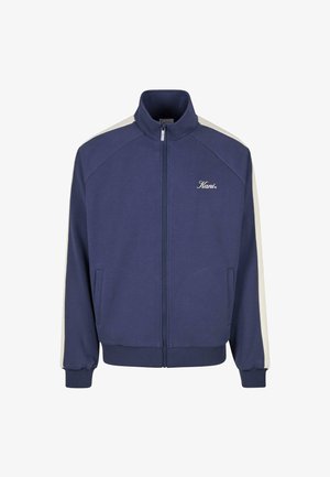 Jūras zilas lidošanas jaka ar rāvējslēdzēju, baltām plecu un piedurkņu svītrām, ribotām aprocēm un apakšmalu, kā arī "Kani" logo kreisajā krūtī.