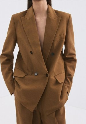 Vrouw draagt een bruin dubbelgeklede blazer met zwarte knopen en bijpassende broek, handen in de zakken, tegen een eenvoudige lichte achtergrond.