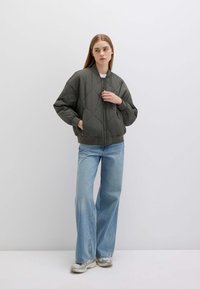 Giacca puffer di colore verde scuro con motivo a rombi, chiusura con cerniera e polsini a costine, abbinata a jeans svasati di colore blu chiaro e sneakers.