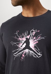 Schwarzes Langarm-T-Shirt mit einem weißen und rosa Grafik eines springenden Figures mit einem Basketball, vor einem explosiven Hintergrund.