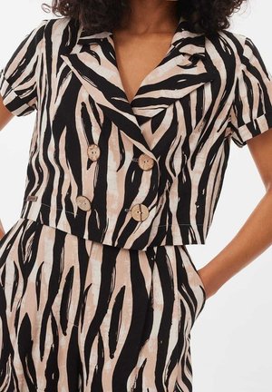 Frau trägt eine kurzärmelige, zweireihige, kurze Jacke und passende hoch taillierte Hosen mit schwarzen und beigen Zebrastrifen.
