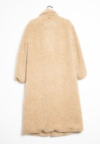 Beige oversized kabát vyrobený z měkkého, huňatého materiálu. Má límec s výstřihem do V a spadlé ramena. Spodní lem je mírně vlnkovaný.