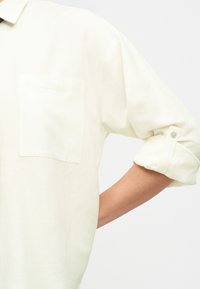 Cămașă albă cu mânecă lungă, suflecată, cu un buzunar frontal pe partea stângă și o bandă cu nasture pe mânecă.