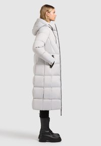 Long, light grey puffer coat with hood, featuring quilted sections, front zipper, and side pockets, paired with black lace-up boots.
