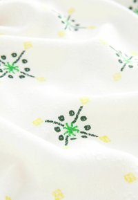 Tela blanca con patrones florales bordados en verde, negro y amarillo, presentada con suaves pliegues y detalles de textura.