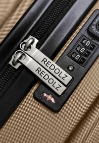 Dos cremalleras plateadas dobles etiquetadas "REDOLZ" aseguradas con un candado de combinación negro sobre una superficie de equipaje texturizada en color beige.