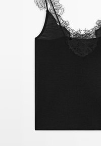 Top nero senza maniche con bordo in pizzo floreale trasparente lungo la scollatura e la parte superiore del petto su uno sfondo bianco.