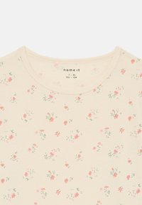 Camiseta de color beige claro con estampado floral y escote redondo, que presenta pequeñas flores rosas y verdes esparcidas por la tela.