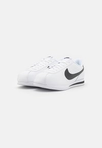 White athletic shoes made of synthetic material, featuring a black Nike swoosh, textured sole, and classic lace-up design.