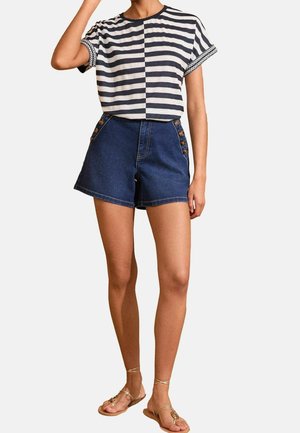 Vrouw draagt een donkerblauw-wit gestreept shirt met korte mouwen, donkergekleurde denim shorts met hoge taille en knopen aan de zijkant, en beige sandalen met bandjes.