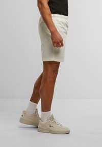 Shorts beige clair avec une taille élastique, des poches latérales et une coupe décontractée, associés à des baskets couleur crème et des chaussettes blanches.