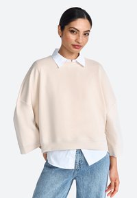 Sudadera de gran tamaño en beige claro con un cuello blanco, corte relajado, hombros caídos y una textura mate. Combinada con una camisa blanca.