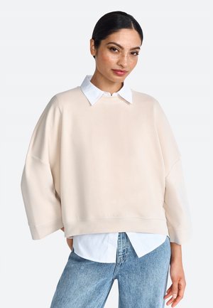 Sudadera de gran tamaño en beige claro con un cuello blanco, corte relajado, hombros caídos y una textura mate. Combinada con una camisa blanca.