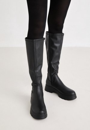 Bottes en cuir noires montantes jusqu'aux genoux avec fermetures éclair latérales portées avec des collants opaques noirs sur un sol clair.