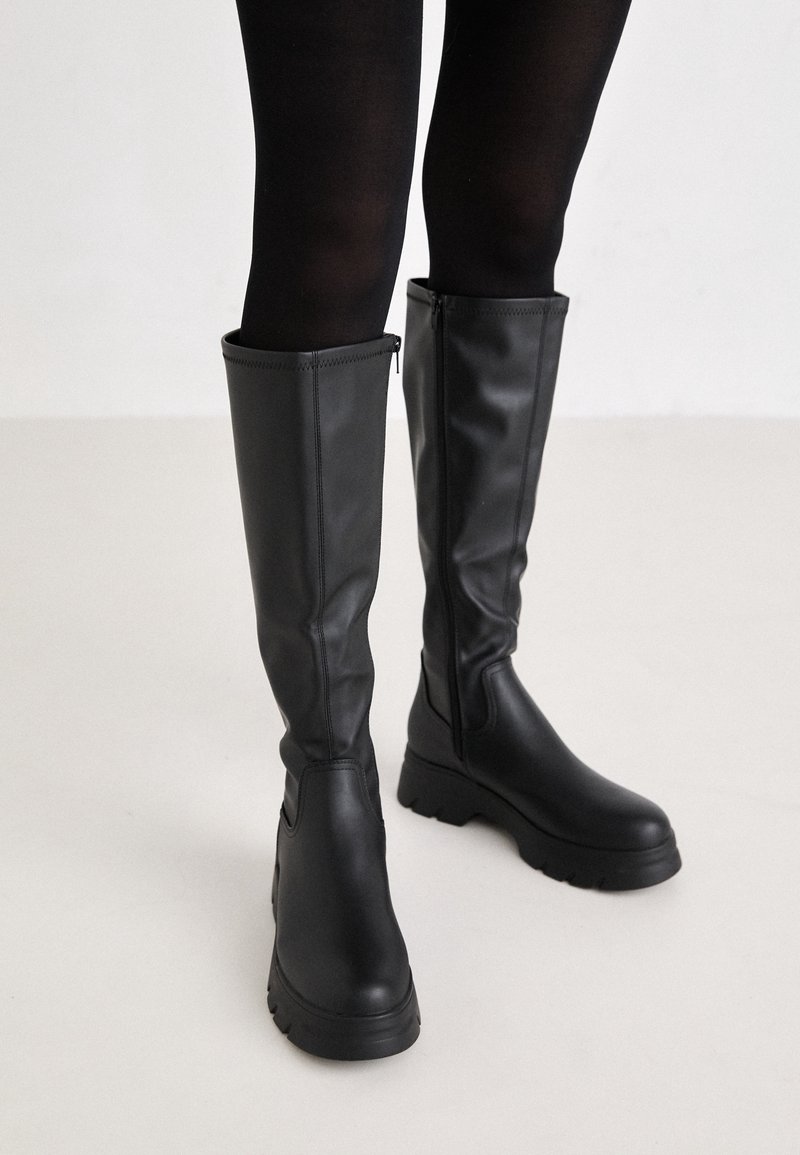 Bottes en cuir noires montantes jusqu'aux genoux avec fermetures éclair latérales portées avec des collants opaques noirs sur un sol clair.