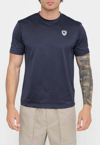 T-shirt blu navy a maniche corte realizzato in un materiale liscio, con una piccola toppa con logo sulla parte superiore sinistra del petto, abbinato a pantaloni beige chiaro.