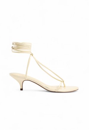 Sandalia beige con tiras, tacón bajo tipo kitten y correas que envuelven el tobillo, diseño minimalista de punta abierta sobre fondo blanco.