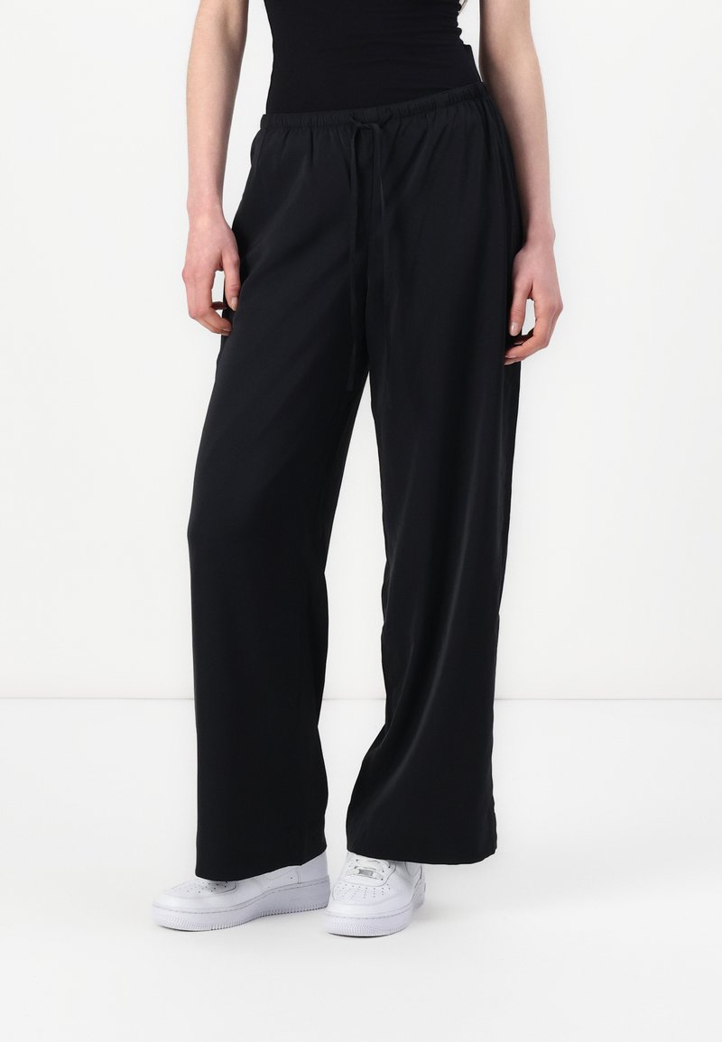 Sorte vide bukser laget av et lett, glatt stoff med elastisk midjebånd og snor, parret med hvite joggesko.